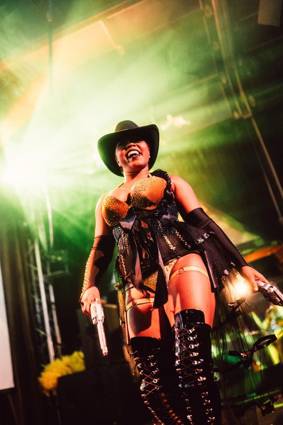 A performer in a black cowboy hat, gold corset and thigh-high lace-up boots throws their head back laughing in green haze