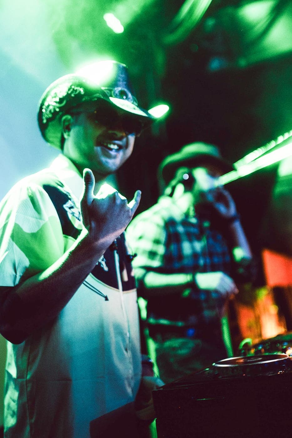 A DJ in a cowboy hat and sunglasses behind the decks in green light, a second performer in plaid beside them