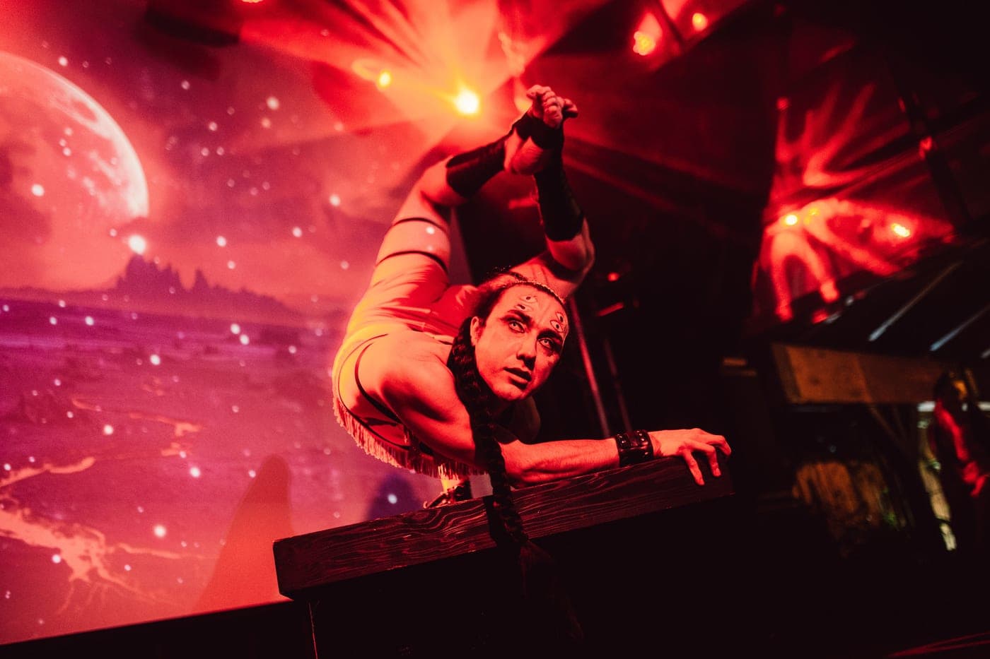 Two performers in deep red light, one balanced low across a wooden block