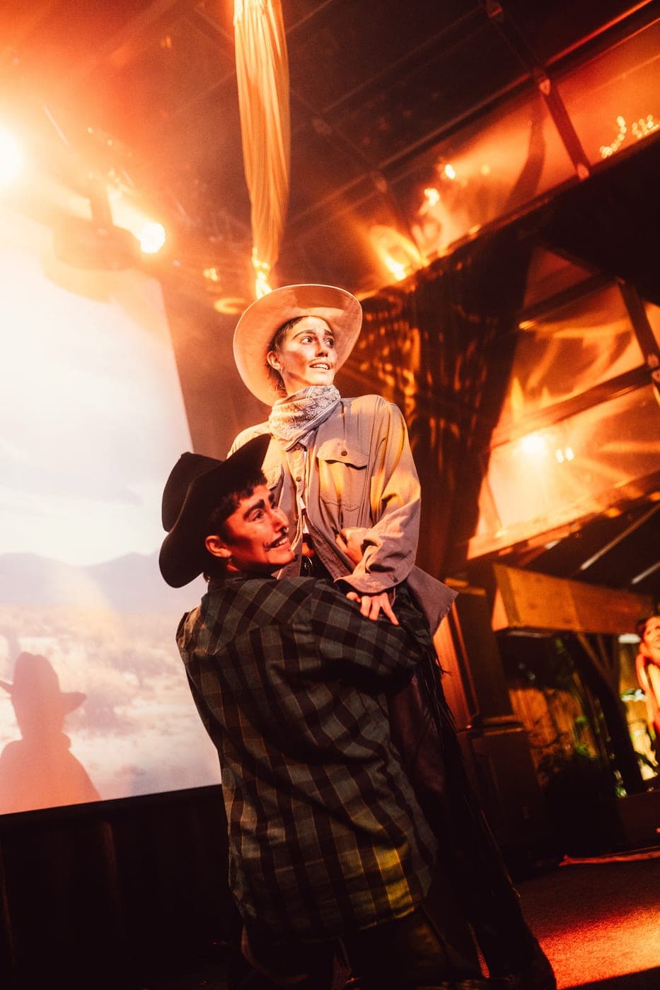 Two performers in cowboy hats hold each other close in front of an orange-lit desert backdrop