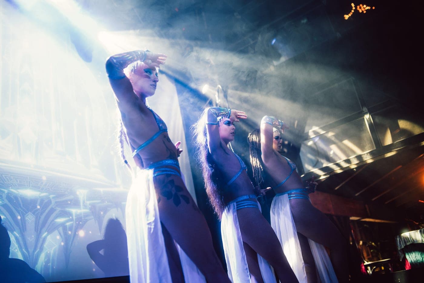 Three performers in silver and white alien costumes raise their arms together under blue stage light